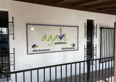 Agave Pediatrics entrance sign in Phoenix, crafted by Arizona Custom Signworks, directs to well and sick entries on a white wall.